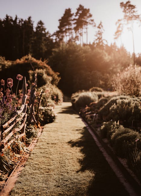 A neatly maintained garden pathway lined with a variety of flowering plants and shrubs on the left side, including purple and pink blossoms, and a row of trimmed bushes and small trees on the right. T