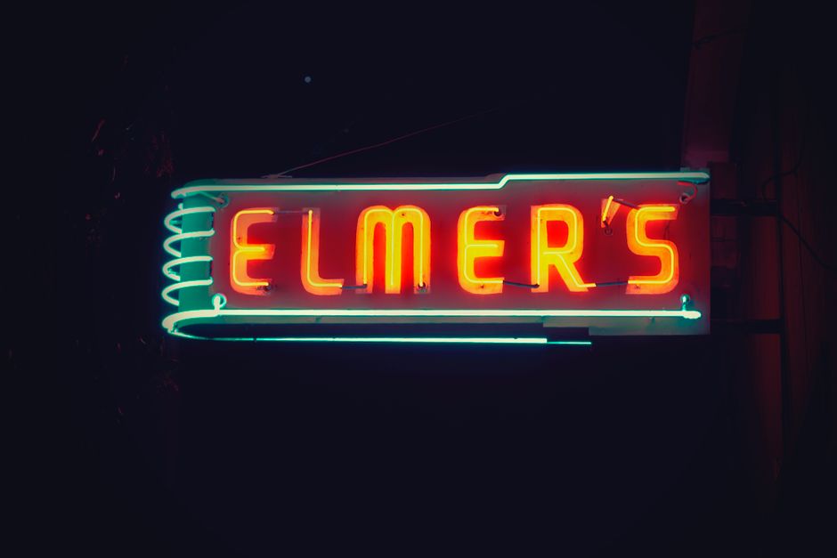 A neon sign displaying the word 'ELMER'S' in bright red letters, outlined by a blue neon border on a dark background. The sign appears to be mounted on an exterior wall, with visible electrical wiring