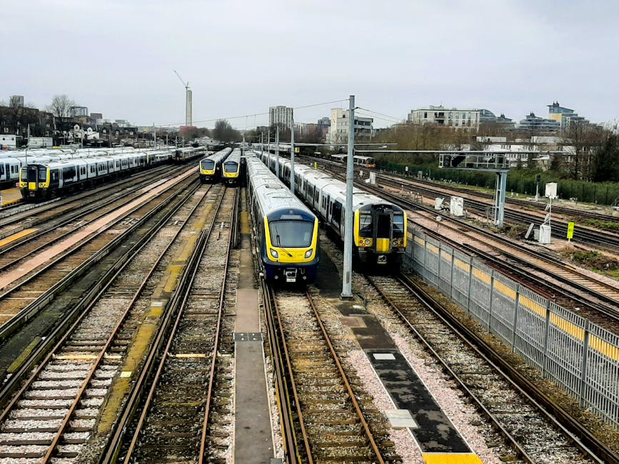 A busy railway yard with multiple parallel train tracks, some with modern yellow and grey commuter trains, parked and ready for departure. The tracks are made of steel rails set on wooden sleepers, wi
